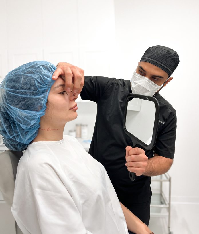 Surgeon in a mask holding a hand mirror beside a patient in a blue surgical cap during a pre-operative consultation
