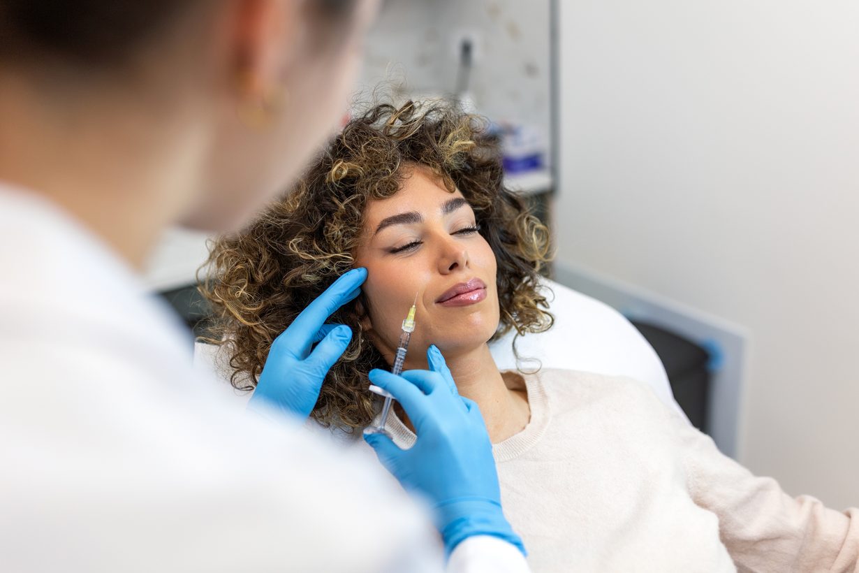 Gloved hands injecting dermal filler into the cheek of a reclining patient